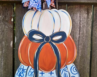 May include: A wooden pumpkin decoration with three stacked sections. The top section is white, the middle is orange, and the bottom is light blue with a floral design. A navy blue bow adorns the center, and a multi-patterned bow tops the pumpkin.
