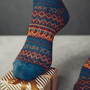 May include: Close-up of a blue sock with a geometric pattern in red and yellow. The sock is resting on a small gift box wrapped in gold and white striped paper, tied with a gold ribbon.