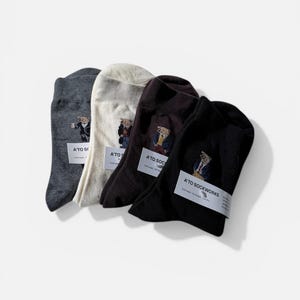 Op de afbeelding: Vier paar sokken in antracietgrijs, crème, donkerbruin en zwart. Elk paar is voorzien van een geborduurde beer in een pak en is vastgezet met een witte papieren band met het label A'TO SOCKWORKS en onderhoudsinstructies in zwarte tekst.