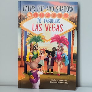 May include: A children's book titled "Tater Tot and Shadow Welcome to Fabulous Las Vegas." The cover features cartoon animal characters in Las Vegas attire, with the iconic "Welcome to Fabulous Las Vegas" sign. The book is written by Laurie Eden and illustrated by Folksnfables.