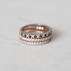 May include: Three stacking rings with a pave setting of small clear and black diamonds. The rings are in different metal finishes: rose gold, white gold, and silver.