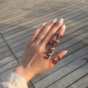 Könnte beinhalten: Eine Hand trägt einen zarten Ring und einen schwarzen und goldenen Statement-Ring mit floralem Design. Der Ring ist mit einer Kette verbunden, die sich um den Finger wickelt.