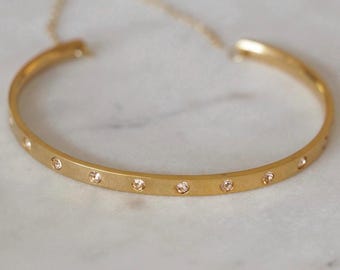 Puede incluir: Un brazalete dorado con pequeñas piedras preciosas transparentes espaciadas uniformemente en la parte delantera. El brazalete tiene un diseño simple y elegante y una delicada cadena. La joya se exhibe sobre una superficie de mármol blanco.