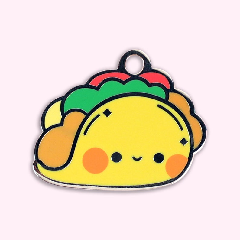 May include: A yellow, cartoon-style taco charm with a smiling face, red and green toppings, and a silver-colored loop at the top. The charm has a black outline and is set against a light pink background.
