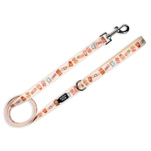 Può includere: Un guinzaglio per cani beige con un motivo colorato di caramelle e altri dolcetti. Il guinzaglio ha un moschettone argentato e un logo Fuzzy Paw nero.