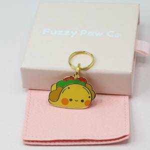 May include: A gold-colored keychain featuring a cartoon taco design with a smiling face, green lettuce, and red tomatoes. The keychain is attached to a gold ring and rests on a pink cloth, with a pink box in the background that reads "Fuzzy Paw Co."