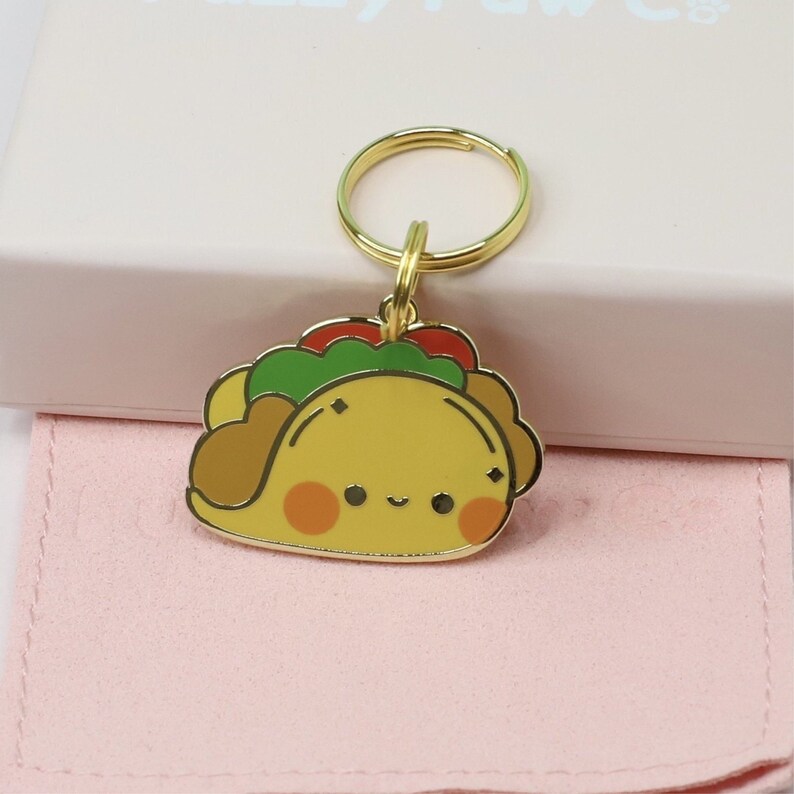 May include: A gold-colored keychain featuring a cartoon taco design. The taco has a yellow shell, brown filling, green lettuce, and red tomatoes. It has a smiling face with orange cheeks. The keychain is on a gold ring.
