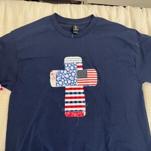 以下が含まれることがあります： アメリカ国旗模様の十字型アップリケが付いたネイビーブルーのTシャツ。袖は折り返されており、赤、白、青の模様の生地が見えます。愛国的なテーマのアパレルアイテム。