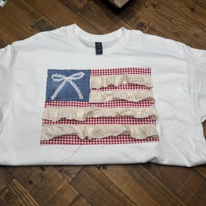 Peut inclure: T-shirt blanc avec un motif patriotique. Le motif présente un carré bleu denim avec un nœud blanc et du vichy rouge et blanc avec des rayures de dentelle crème, ressemblant au drapeau américain.