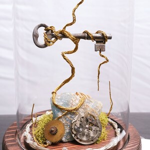 Peut inclure: Un dôme en verre avec une base en bois contient une sculpture fantaisiste d'inspiration steampunk. Une clé en argent est enveloppée de fil doré, et le fil s'étend vers le haut pour former une structure arborescente avec des engrenages et des pièces d'horlogerie. La base est recouverte de mousse et d'une roche bleu-vert.