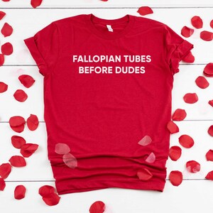 May include: Red t-shirt with white text that reads "Fallopian Tubes Before Dudes". The shirt is surrounded by red rose petals on a white wooden background.