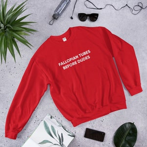 May include: A bright red sweatshirt with the text "FALLOPIAN TUBES BEFORE DUDES" in white block letters. The sweatshirt is displayed on a neutral surface with other accessories, including sunglasses and a water bottle.