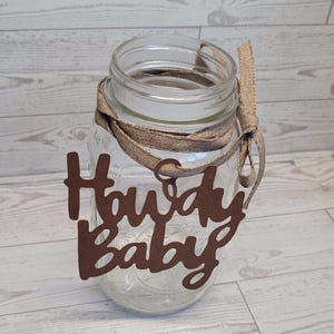 Op de afbeelding: Een doorzichtige glazen mason jar met een bruin uitgesneden bordje met de tekst Howdy Baby. Het bordje is aan de hals van de pot bevestigd met een beige jute lint dat in een strik is geknoopt. De pot heeft een gladde rand en een cilindrisch lichaam.