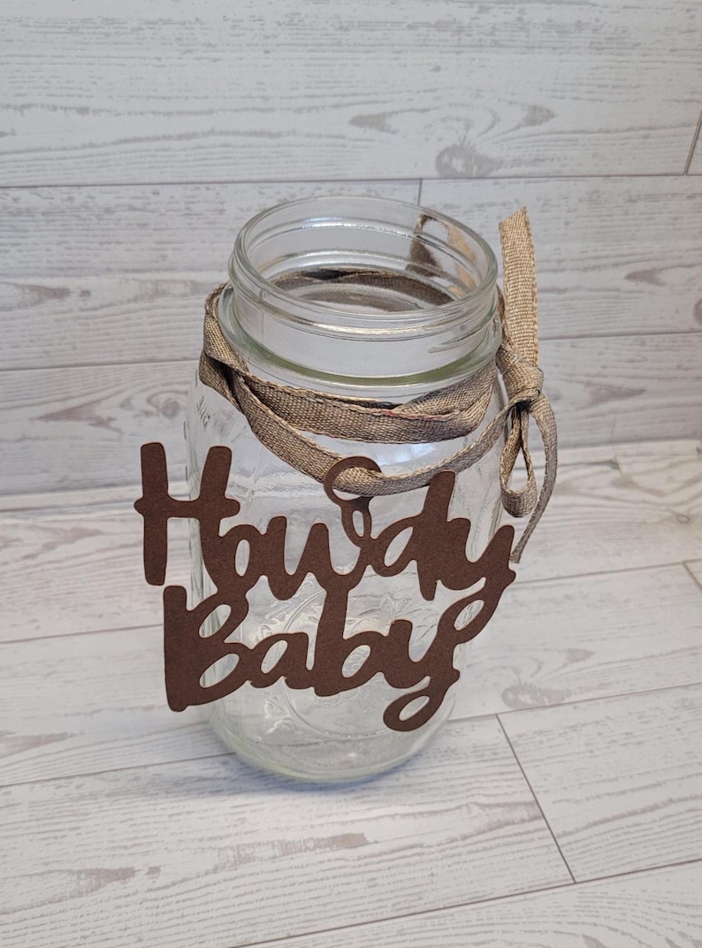 Op de afbeelding: Een doorzichtige glazen mason jar met een bruin uitgesneden bordje met de tekst Howdy Baby. Het bordje is aan de hals van de pot bevestigd met een beige jute lint dat in een strik is geknoopt. De pot heeft een gladde rand en een cilindrisch lichaam.