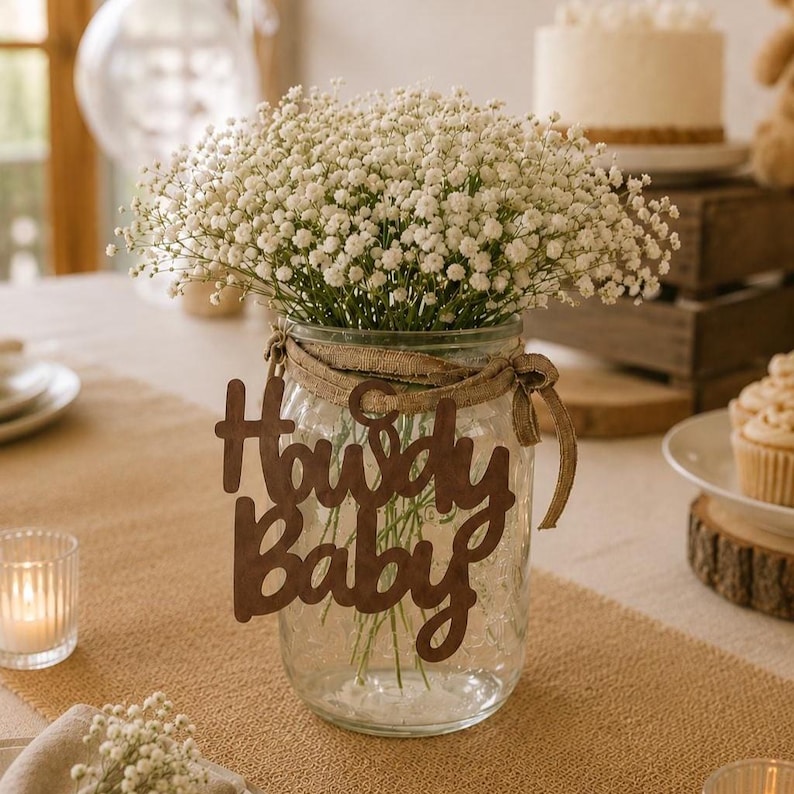 Op de afbeelding: Een doorzichtige glazen mason jar gevuld met witte gipskruidbloemen. Een bruin houten uitsnede met de tekst Howdy Baby is aan de pot bevestigd met een beige jute lint. Het arrangement wordt gepresenteerd als een decoratief middelpunt.