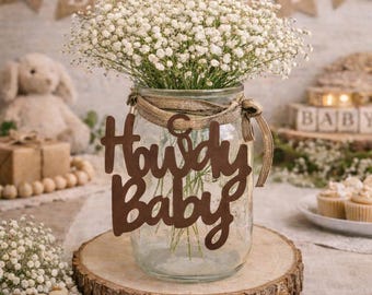 Op de afbeelding: Een doorzichtige glazen pot als tafelstuk gevuld met witte gipskruidbloemen. De pot is voorzien van een bruin uitgesneden bordje met de tekst Howdy Baby en is vastgebonden met een gestructureerd beige lint op een ronde houten schijf.