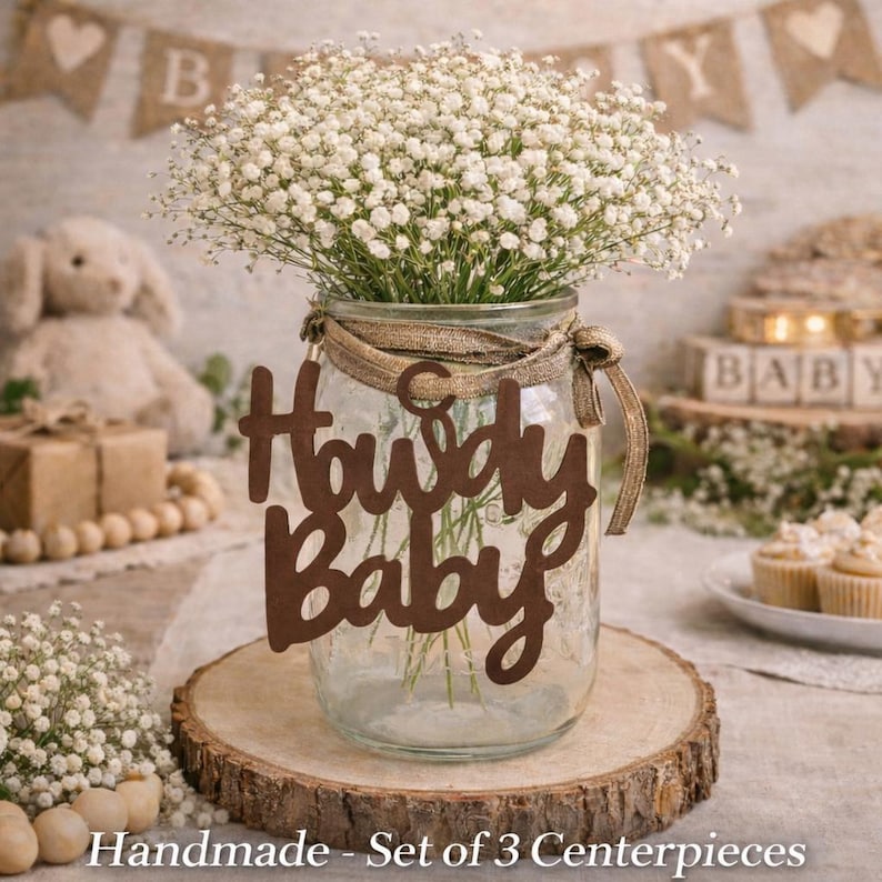 Op de afbeelding: Een doorzichtige glazen pot als tafelstuk gevuld met witte gipskruidbloemen. De pot is voorzien van een bruin uitgesneden bordje met de tekst Howdy Baby en is vastgebonden met een gestructureerd beige lint op een ronde houten schijf.
