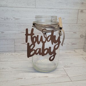 Op de afbeelding: Een doorzichtige glazen mason jar met een bruin uitgesneden bordje met de tekst Howdy Baby in een cursief lettertype. Het bordje is aan de hals van de pot bevestigd met een beige stoffen lint dat in een strik is geknoopt.