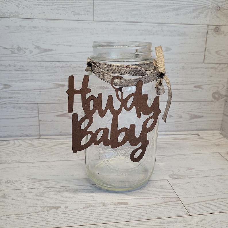 Op de afbeelding: Een doorzichtige glazen mason jar met een bruin uitgesneden bordje met de tekst Howdy Baby in een cursief lettertype. Het bordje is aan de hals van de pot bevestigd met een beige stoffen lint dat in een strik is geknoopt.