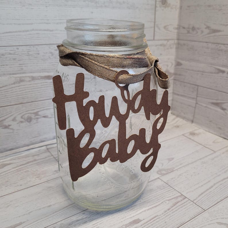Op de afbeelding: Een doorzichtige glazen weckpot met een bruin uitgesneden bordje met de tekst Howdy Baby, bevestigd aan de hals met een bruin stoffen lint. De pot heeft een wijde opening en een schroefdraad, wat een transparante houder voor decoratie vormt.