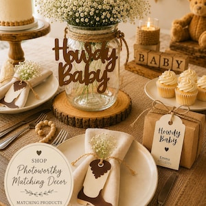 Op de afbeelding: Een glazen pot als middelpunt gevuld met witte gipskruidbloemen, voorzien van een bruin uitgesneden bordje met de tekst Howdy Baby. De pot staat op een houten schijf, omringd door borden, servetten met laarsvormige labels en een geschenkdoos.