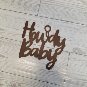 Op de afbeelding: Een bruin, lasergesneden houten bord met de tekst Howdy Baby in een cursief lettertype. Het bord bevat een kleine cirkelvormige uitsparing in het midden bovenaan om op te hangen, met een matte afwerking en strakke, donkergekleurde randen.