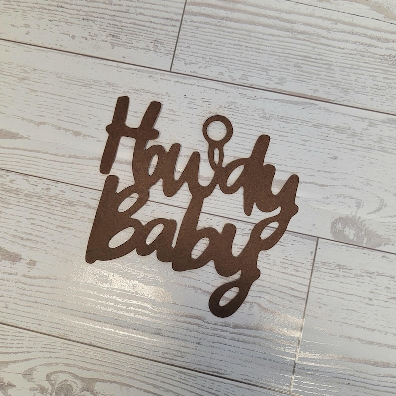 Op de afbeelding: Een bruin, lasergesneden houten bord met de tekst Howdy Baby in een cursief lettertype. Het bord bevat een kleine cirkelvormige uitsparing in het midden bovenaan om op te hangen, met een matte afwerking en strakke, donkergekleurde randen.