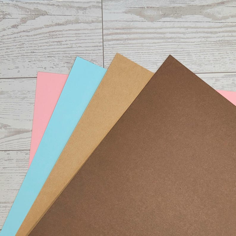 Op de afbeelding: Een stapel van vier rechthoekige vellen kartonpapier die in een waaierpatroon zijn gerangschikt. De vellen zijn gekleurd in tinten lichtroze, lichtblauw, beige en donkerbruin. Elk vel heeft een gladde, matte afwerking en strakke randen.