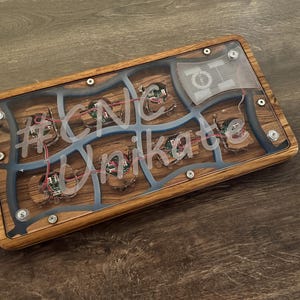 May include: A rectangular wooden box with a clear top, featuring internal components and the text "#CNC Unikate". The box has a wood grain pattern, blue accents, and visible wiring. The design incorporates a mix of natural and technological elements.