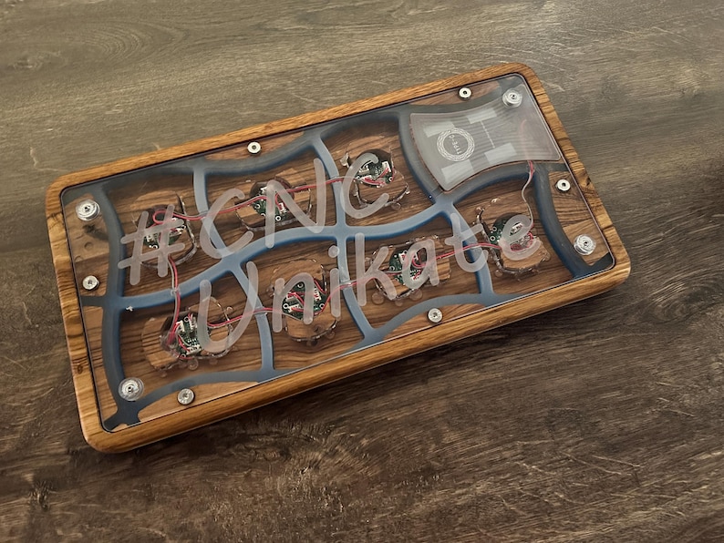 May include: A rectangular wooden box with a clear top, featuring internal components and the text "#CNC Unikate". The box has a wood grain pattern, blue accents, and visible wiring. The design incorporates a mix of natural and technological elements.