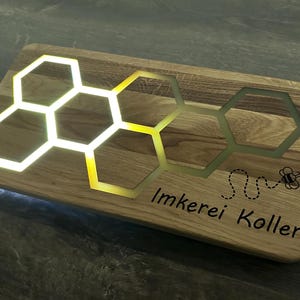 K&ouml;nnte beinhalten: Ein Holzbrett mit einem Wabenmuster und dem Text "Imkerei Koller" sowie einer Bienenillustration. Die Wabe ist mit wei&szlig;em Licht beleuchtet.