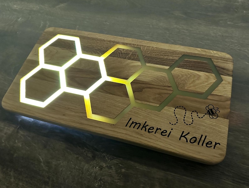 K&ouml;nnte beinhalten: Ein Holzbrett mit einem Wabenmuster und dem Text "Imkerei Koller" sowie einer Bienenillustration. Die Wabe ist mit wei&szlig;em Licht beleuchtet.