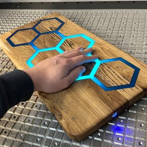 K&ouml;nnte beinhalten: Ein Schneidebrett aus Holz mit einer blauen Epoxidharz-Einlage in Form eines Wabenmusters. Das Epoxidharz leuchtet blau und das Brett wird von unten beleuchtet.