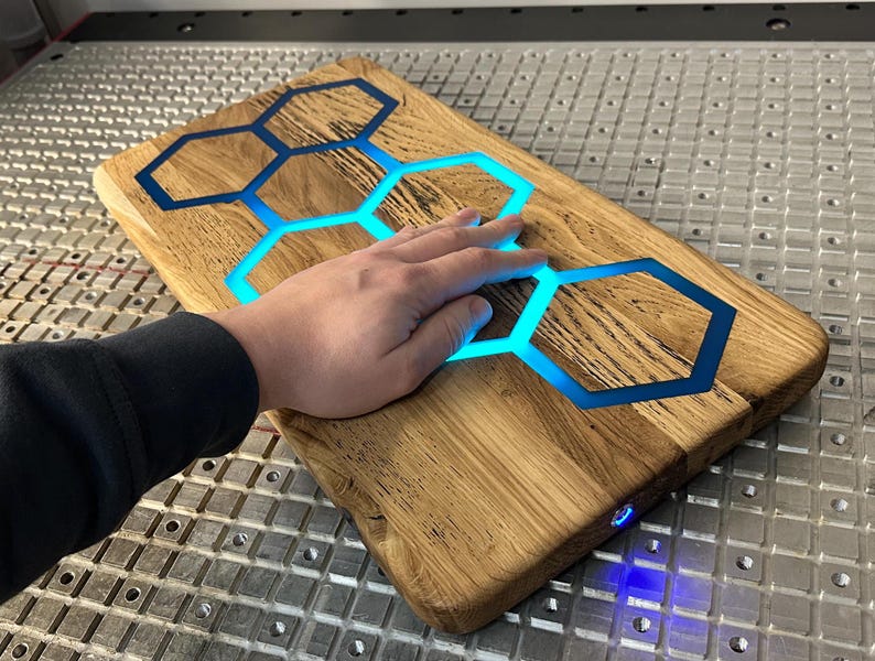 K&ouml;nnte beinhalten: Ein Schneidebrett aus Holz mit einer blauen Epoxidharz-Einlage in Form eines Wabenmusters. Das Epoxidharz leuchtet blau und das Brett wird von unten beleuchtet.