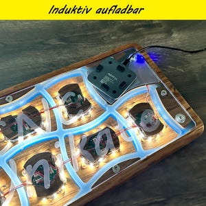 May include: A wooden base with a blue illuminated design and the name "Nika" in white letters. The base has a clear acrylic top and a black charging pad. The text "Induktiv aufladbar" is in the top yellow bar.