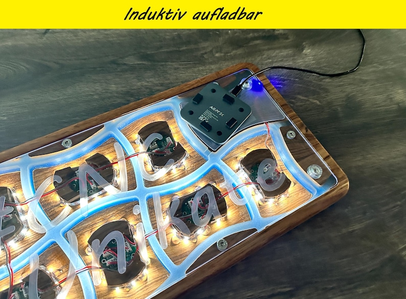 May include: A wooden base with a blue illuminated design and the name "Nika" in white letters. The base has a clear acrylic top and a black charging pad. The text "Induktiv aufladbar" is in the top yellow bar.