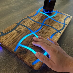 May include: Wooden serving board with a unique design. The board features a network of blue glowing resin, creating an eye-catching pattern. A dark bottle sits on the board. The board is on a wooden surface.