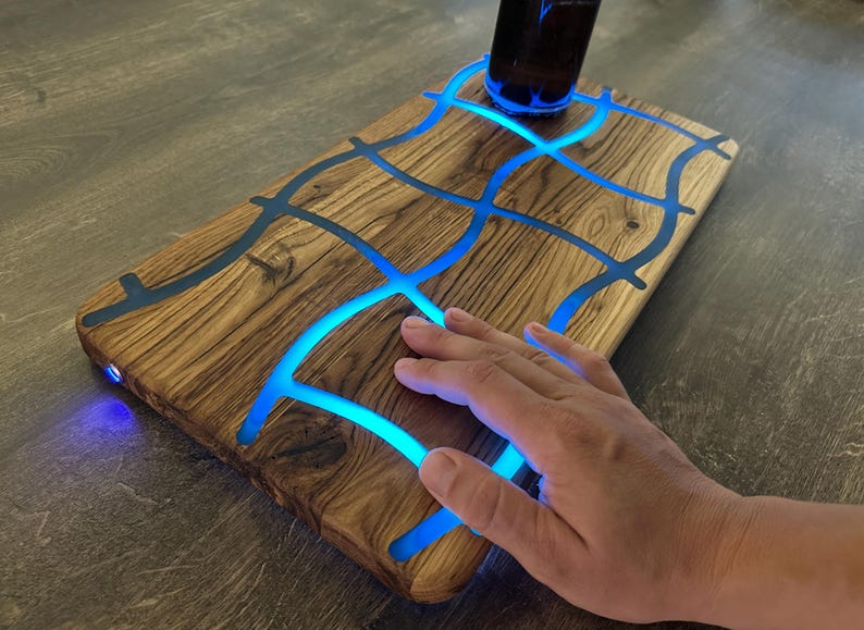 May include: Wooden serving board with a unique design. The board features a network of blue glowing resin, creating an eye-catching pattern. A dark bottle sits on the board. The board is on a wooden surface.
