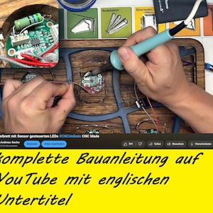 May include: A person soldering wires to a circuit board, part of a wooden serving board with LED lights. The image includes text in German, advertising a complete building guide on YouTube with English subtitles.
