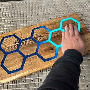 K&ouml;nnte beinhalten: Ein Schneidebrett aus Holz mit einer blauen Epoxidharzeinlage in Form eines Wabenmusters. Das Brett wird von unten beleuchtet, wodurch das Harz leuchtet.