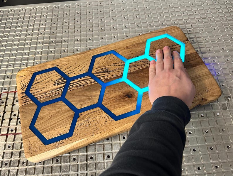 K&ouml;nnte beinhalten: Ein Schneidebrett aus Holz mit einer blauen Epoxidharzeinlage in Form eines Wabenmusters. Das Brett wird von unten beleuchtet, wodurch das Harz leuchtet.