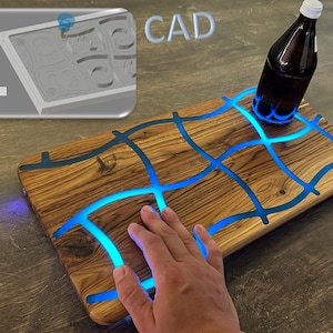 May include: A wooden serving board with blue glowing resin accents. A brown bottle with a white cap sits on the board. The word "CAD" is in the background. The board is rectangular with a hand touching the surface.