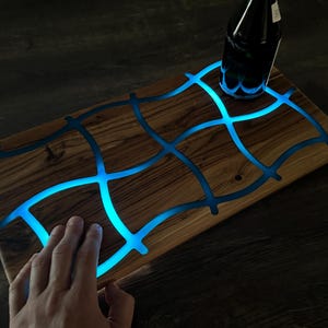 May include: Wooden serving board with a unique design. The board features a grid pattern of blue resin that glows with a soft blue light. A dark bottle sits on the board. The board is rectangular and has a natural wood grain.