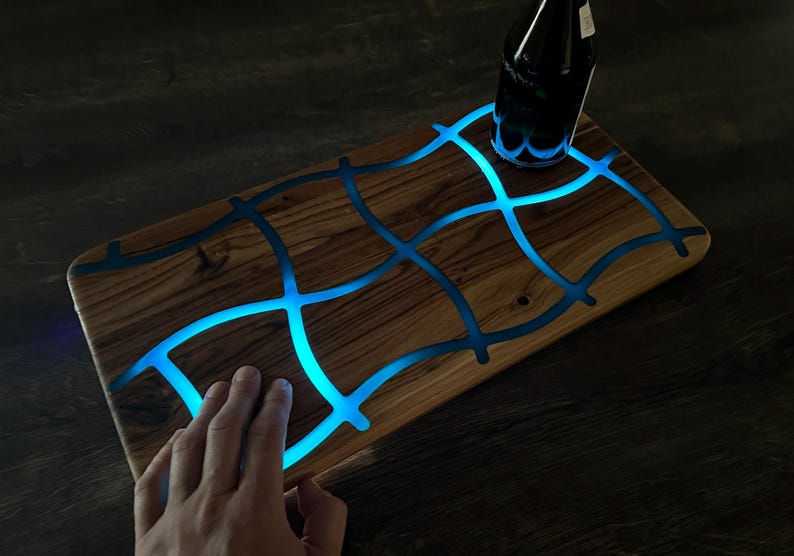 May include: Wooden serving board with a unique design. The board features a grid pattern of blue resin that glows with a soft blue light. A dark bottle sits on the board. The board is rectangular and has a natural wood grain.