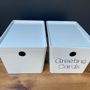 May include: Two white plastic storage boxes with lids. The box on the right has the words "Greeting Cards" written on the front in blue.
