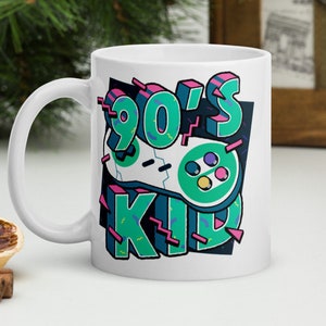 May include: White ceramic mug with a retro 90's design. The mug features the text "90's KID" in teal, pink, and black, along with a stylized video game controller graphic. The mug has a curved handle.