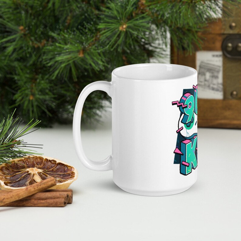 May include: White ceramic mug with a large handle. The mug features the text "90s Kid" in a colorful, stylized font with teal, pink, and black accents. The mug is set against a background with greenery and a wooden chest.