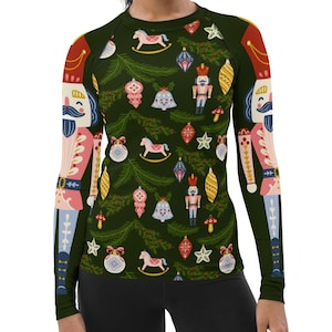Peut inclure: Un t-shirt à manches longues avec un fond vert et un motif répété de casse-noix, d'ornements et de chevaux à bascule. Les casse-noix portent des chapeaux rouges et ont des accents roses et bleus. Les ornements sont rouges, bleus, verts et jaunes. Les chevaux à bascule sont roses.
