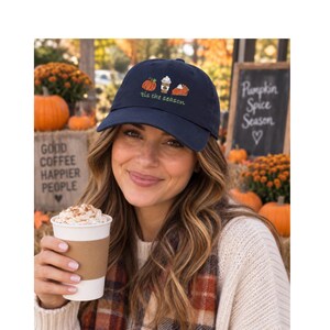 May include: A navy blue baseball cap featuring an embroidered design of a pumpkin, a coffee cup, and a slice of pie, with the text 'tis the season' in green script lettering centered below the icons on the front panel of the hat.