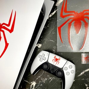 May include: A white PlayStation 5 console and controller with a red spider logo. The image also shows a sheet of red spider decals and two smaller spider decals. The items are on a black and gray marble surface.